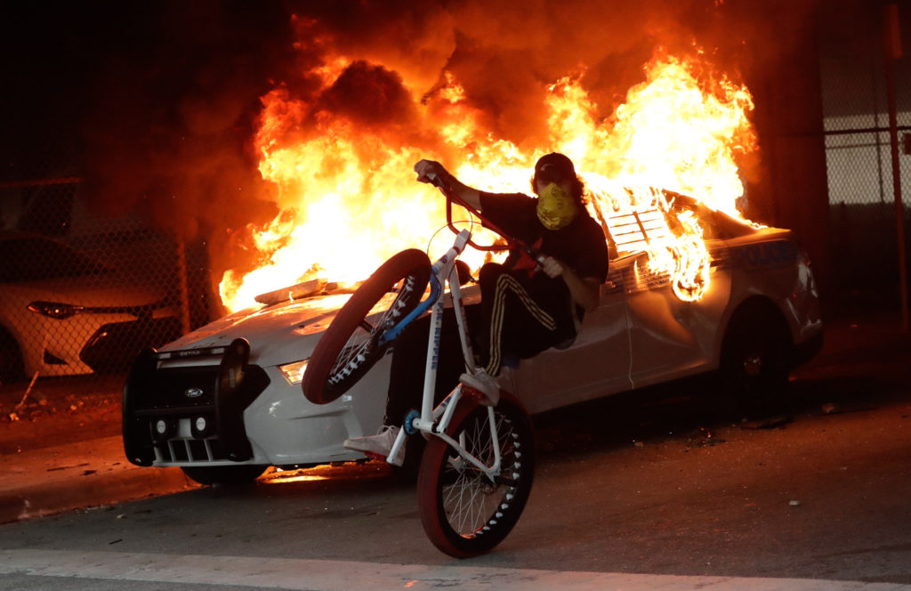 Man popping a wheelie on a bicycle in front of a burning police car